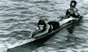 The standard surf lesson assisted by a push from the instructor; the beach boy is also helping to learn balance on the board.  The beach boys were true “ambassadors of aloha” and the first lifeguards on Waikiki Beach.